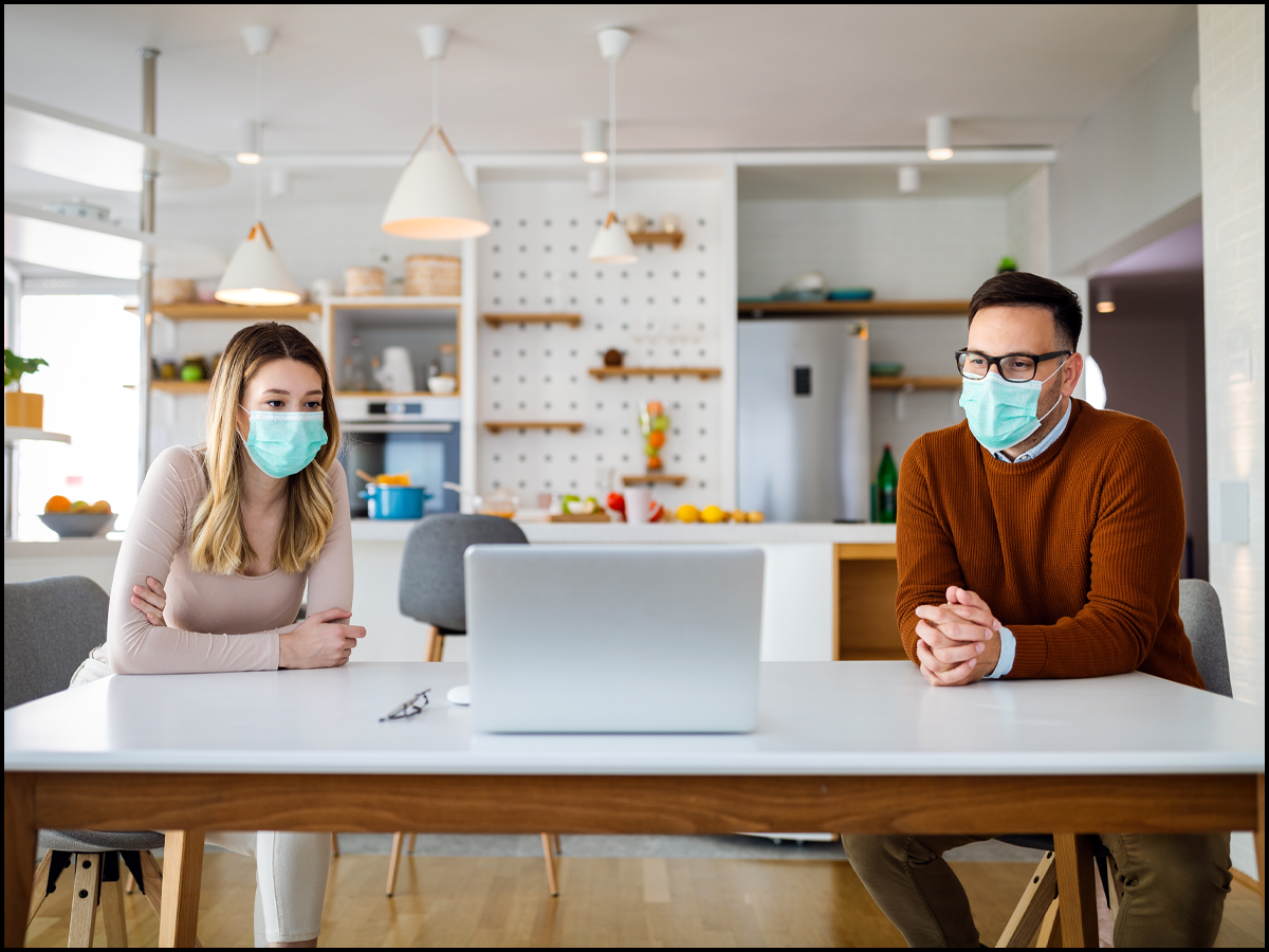 Two people with masks on sitting opposite sides of a table looking at a laptop